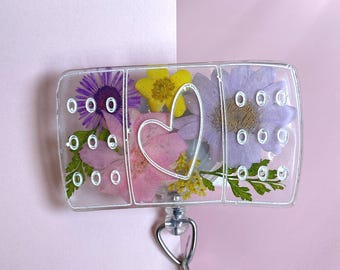 May include: A clear plastic badge holder shaped like a band-aid with pressed flowers inside. The flowers are purple, pink, and yellow. A white heart is in the center of the badge holder.