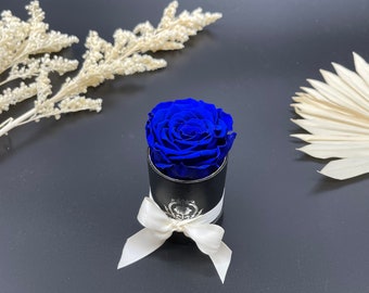 May include: A single blue rose preserved in a black cylinder box with a white ribbon. The rose is in a small box with a silver logo.