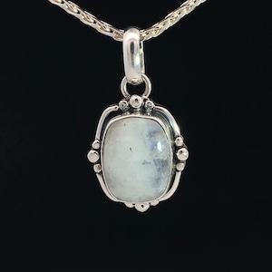 May include: A silver pendant with a white moonstone set in a silver bezel. The pendant is oval shaped and has a detailed design around the stone.