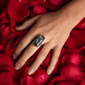 May include: A silver ring with a rectangular blue labradorite gemstone set in a bezel setting. The ring is on a hand resting on a bed of red rose petals.