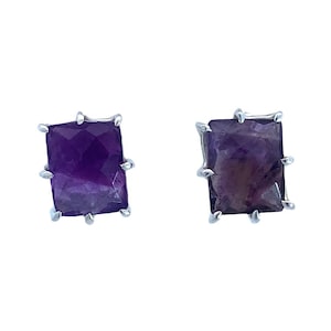 May include: A pair of rectangular amethyst stud earrings. Each earring features a faceted, deep purple gemstone set in a silver-colored metal frame. The frame has a textured, prong-like design around the stone.
