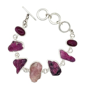 May include: A silver bracelet with a toggle clasp features a variety of pink and purple gemstones. The bracelet is made with a chain link design and has a polished finish.