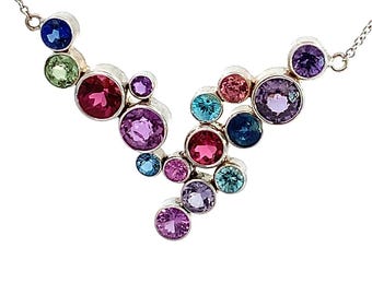 May include: A silver necklace featuring a cluster of colorful gemstones in shades of blue, green, pink, and purple. The gemstones are round and set in individual silver bezels, creating a vibrant and eye-catching design. The necklace has a delicate chain.