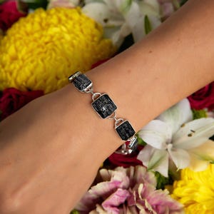 May include: A silver bracelet featuring rectangular black stones. The bracelet is connected by silver links and is displayed on a person's arm against a backdrop of colorful flowers.