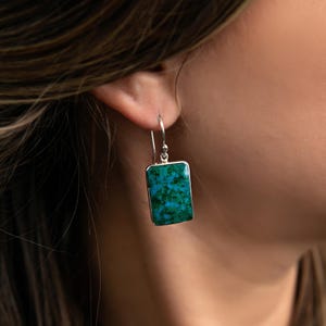May include: Silver-tone rectangular earrings with a vibrant turquoise and green stone inlay. The earrings have a simple hook design and are shown worn on an ear.