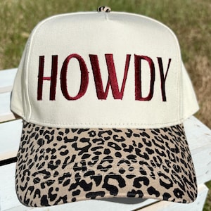 Könnte beinhalten: Cremefarbene Baseballkappe mit dem Wort "HOWDY" in Burgunderrot gestickt. Der Schirm hat einen braunen und schwarzen Leopardenmuster. Ein stilvolles Accessoire für den lässigen Gebrauch.
