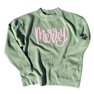 May include: A sage green sweatshirt with the word "Merry" embroidered in pink script across the chest. The sweatshirt has a mock neck and long sleeves, perfect for the holiday season.