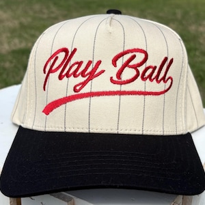 Może przedstawiać: Kremowa czapka bejsbolowa z czarnymi prążkami i czarnym daszkiem. Napis "Play Ball" jest wyszyty czerwoną czcionką z przodu. Czapka ma klasyczny, strukturalny design.