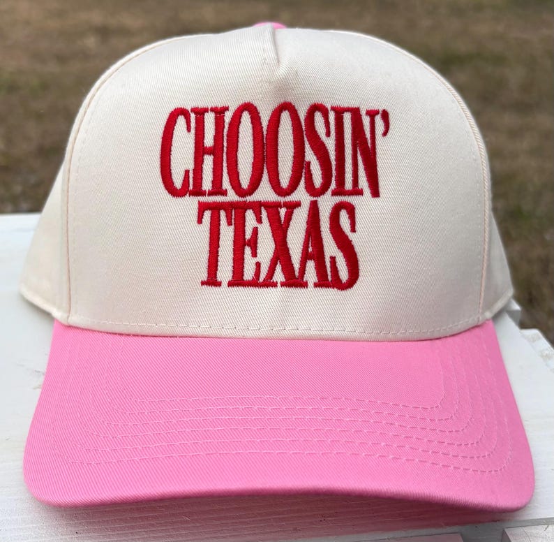 Puede incluir: Gorra de b&eacute;isbol con corona color crema y visera rosa. Las palabras "CHOOSIN' TEXAS" est&aacute;n bordadas en rojo en la parte delantera. La gorra tiene una visera curva y un interior rosa.