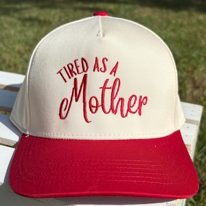 Può includere: Cappellino da baseball color crema con visiera e bottone rossi. La scritta "TIRED AS A Mother" è ricamata in rosso sul davanti. Il cappello è probabilmente realizzato in un tessuto resistente, adatto all'uso quotidiano.
