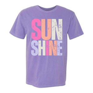 May include: A lilac-coloured t-shirt with short sleeves, featuring the word "SUNSHINE" in a distressed, multi-coloured font. The letters are pink, orange, white, and light blue.