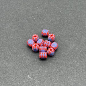 May include: A collection of ten small, barrel-shaped beads. Each bead features a vibrant red base color with a contrasting blue web-like pattern printed across the surface. The beads are arranged in a loose cluster, each showing a central hole.