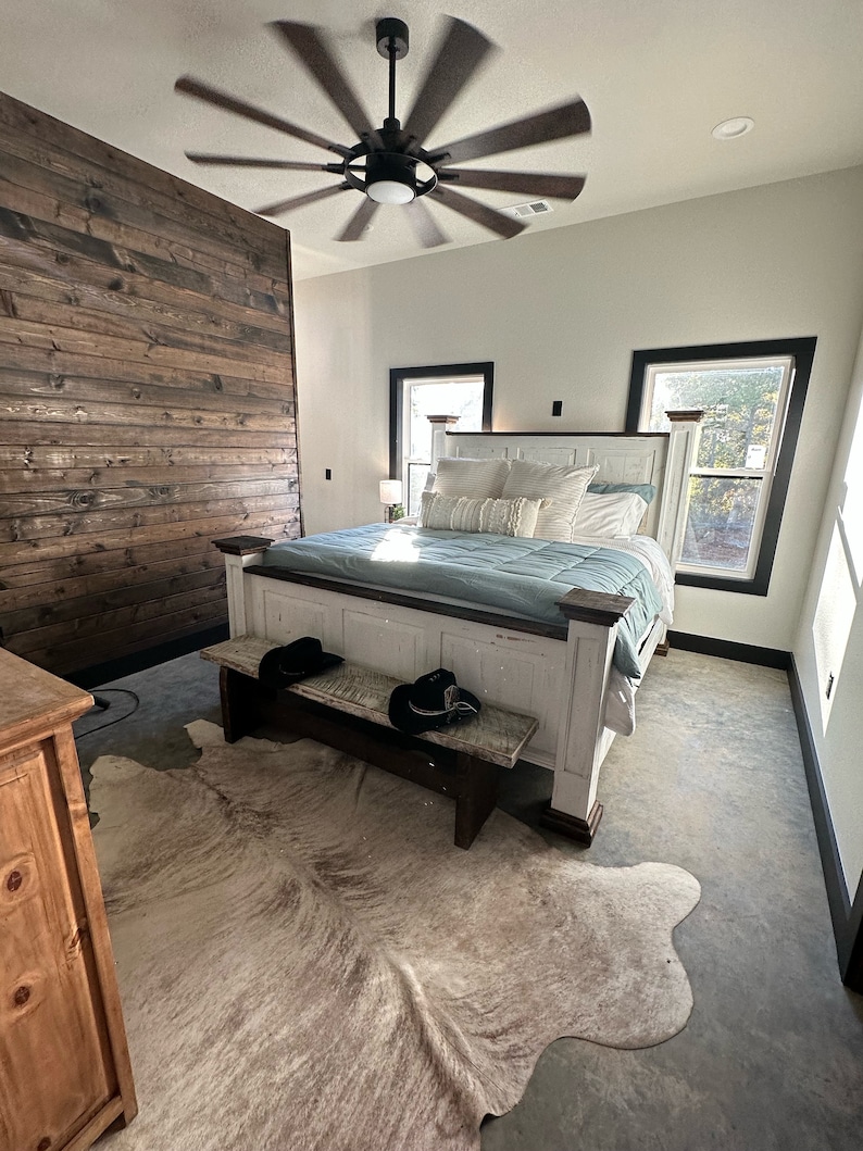 May include: A white bedroom with a large bed, a cowhide rug, and a wooden bench. The bed has a white headboard and a blue blanket. The room has a ceiling fan and two windows with black frames.