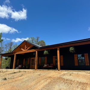 May include: A black house with a large front porch and wooden beams. The house has a black roof and brown wooden trim. There are hanging plants on the porch.