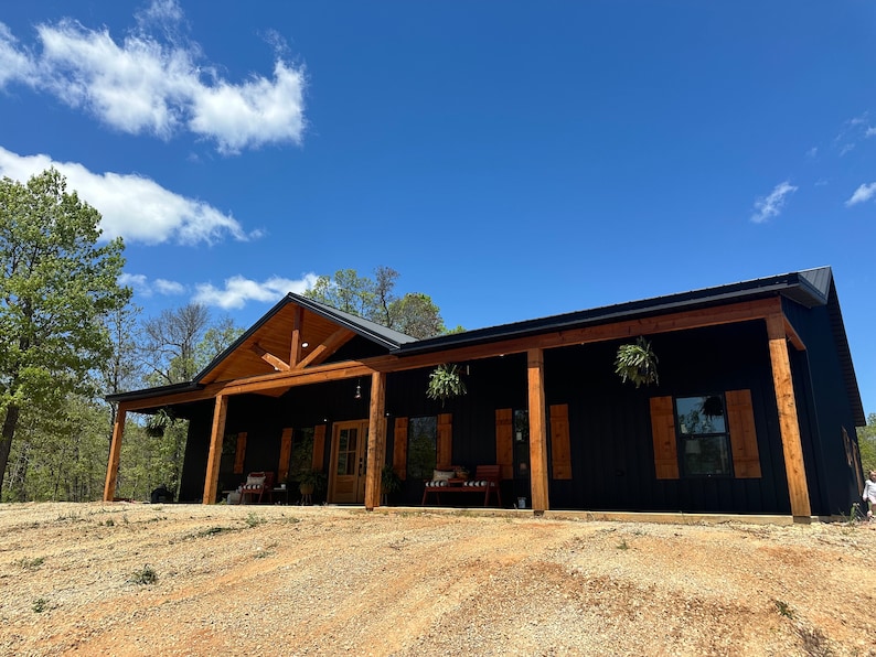 May include: A black house with a large front porch and wooden beams. The house has a black roof and brown wooden trim. There are hanging plants on the porch.
