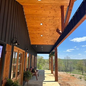 May include: A black metal building with a wooden porch and a view of a wooded area. The porch has a wooden ceiling with recessed lighting and a wooden floor. There are two wooden benches on the porch and a potted plant.
