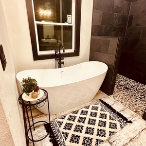 May include: A white freestanding bathtub with a black faucet and a small black metal side table with a potted plant on top. The bathroom has black tile walls and a black and white patterned rug on the floor.
