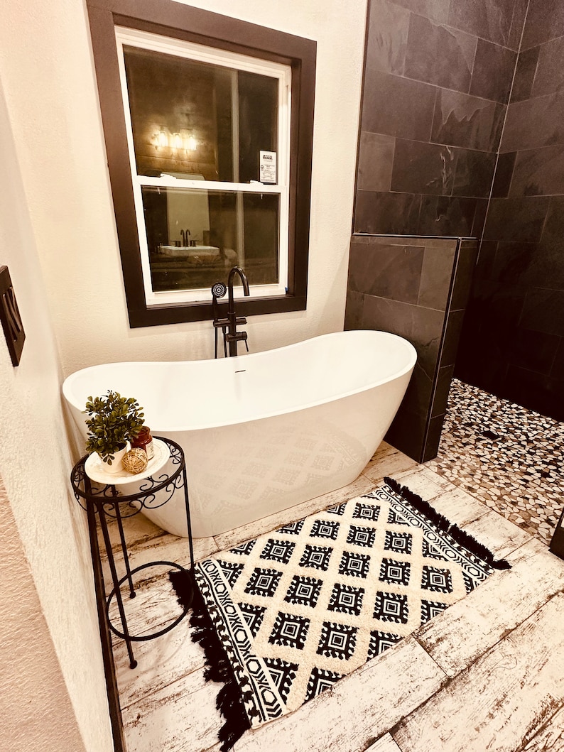 May include: A white freestanding bathtub with a black faucet and a small black metal side table with a potted plant on top. The bathroom has black tile walls and a black and white patterned rug on the floor.