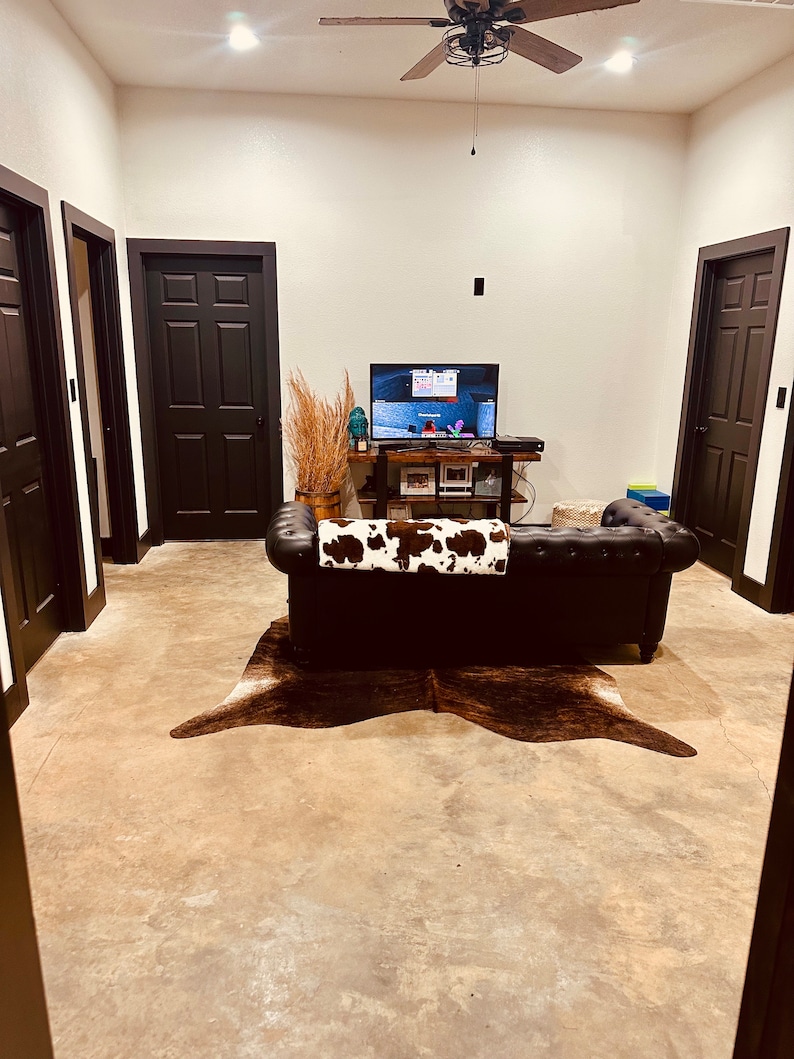May include: A living room with a brown leather couch, a cowhide rug, and a flat screen TV on a wooden shelf. There are three black doors in the room, and a ceiling fan with wooden blades.