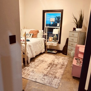 May include: A bedroom with a white bed, a white rug, a pink play kitchen, and a dresser with a plant on top. The bed has a white headboard and a white comforter. The rug is a light brown color with a white pattern. The play kitchen is a light pink color with a black stovetop and a silver sink. The dresser is a light blue color with a white top and four drawers. The plant is a green plant with long, thin leaves.