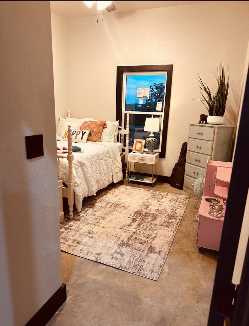 May include: A bedroom with a white bed, a white rug, a pink play kitchen, and a dresser with a plant on top. The bed has a white headboard and a white comforter. The rug is a light brown color with a white pattern. The play kitchen is a light pink color with a black stovetop and a silver sink. The dresser is a light blue color with a white top and four drawers. The plant is a green plant with long, thin leaves.