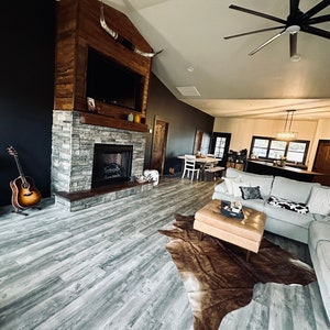 May include: A living room with a fireplace, a large cowhide rug, and a ceiling fan. The fireplace is made of stone and has a wooden mantel. The rug is brown and white and is placed in front of a gray couch. The ceiling fan has wooden blades and is hanging from the ceiling.