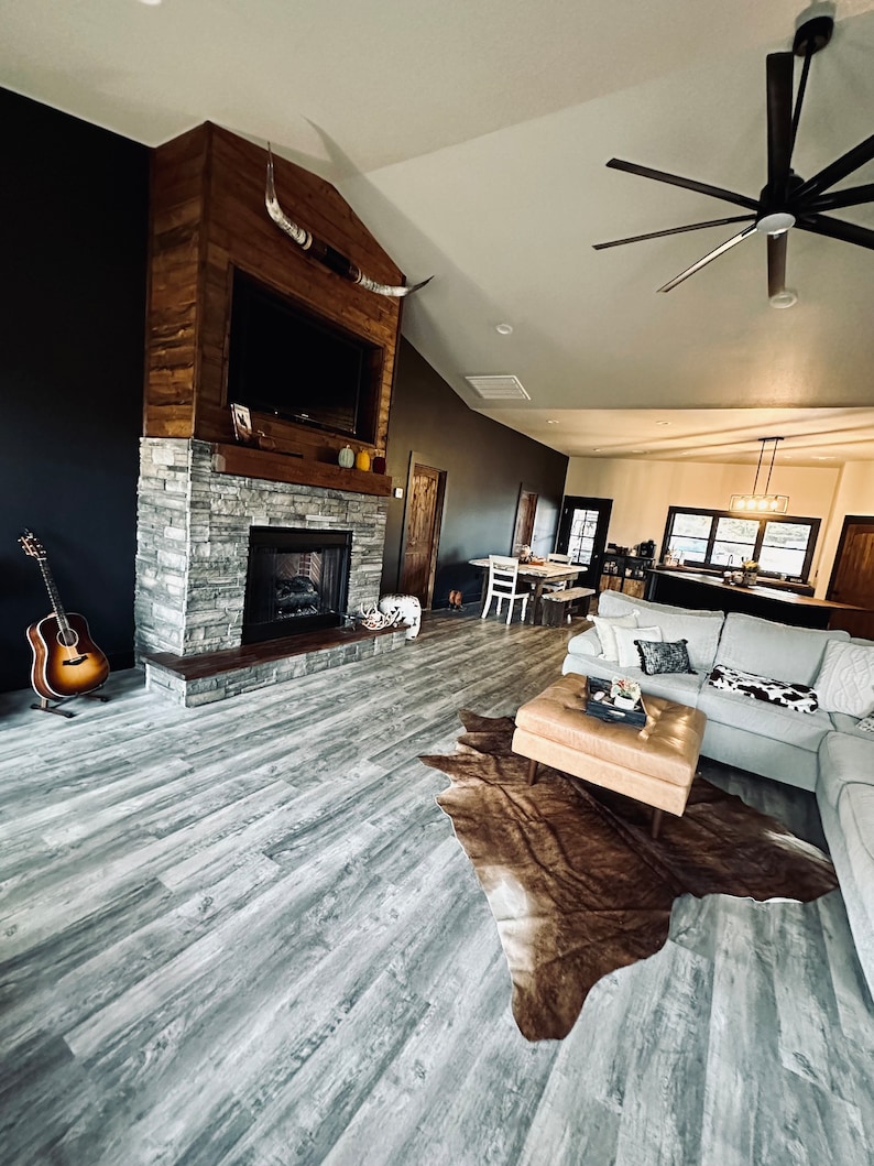 May include: A living room with a fireplace, a large cowhide rug, and a ceiling fan. The fireplace is made of stone and has a wooden mantel. The rug is brown and white and is placed in front of a gray couch. The ceiling fan has wooden blades and is hanging from the ceiling.