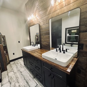 May include: A modern bathroom vanity with two white porcelain sinks, black faucets, and a wood countertop. The vanity is made of black cabinets with brushed nickel hardware. The bathroom features a wood plank wall and a large mirror.