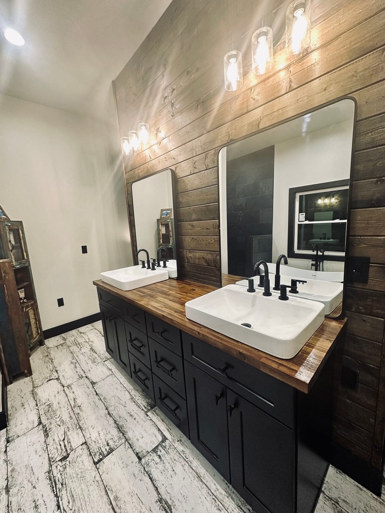 May include: A modern bathroom vanity with two white porcelain sinks, black faucets, and a wood countertop. The vanity is made of black cabinets with brushed nickel hardware. The bathroom features a wood plank wall and a large mirror.