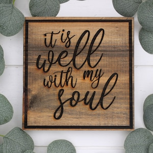 May include: A wooden sign with the text "it is well with my soul" in black lettering. The sign has a dark brown wood grain and a black frame.
