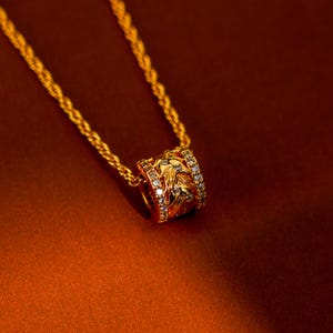 Op de afbeelding: Een goudkleurige ketting met een hanger. De hanger heeft een cilindervorm met ingewikkelde bloemmotieven en kleine, heldere edelstenen. De ketting is ook goudkleurig, de achtergrond is een warm, roodbruin.