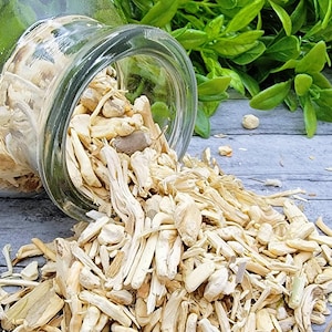 May include: A clear glass jar tipped over, spilling light-colored, dried wood pieces onto a gray wooden surface. Green foliage is visible in the background. The wood pieces vary in size and shape.