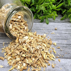 May include: A glass jar filled with dried, light brown, and beige herbal roots, spilling out onto a gray wooden surface.