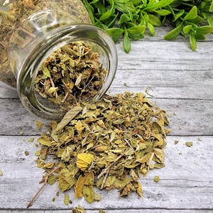 May include: A glass jar with a lid is tipped over, spilling dried green and yellow herbs onto a light gray wooden surface.