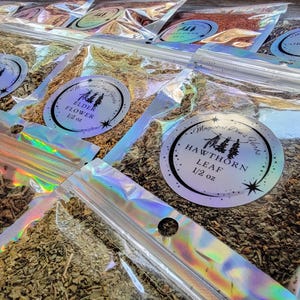 May include: A variety of dried herbs and flowers packaged in clear plastic bags with labels. The labels are white with black text and feature a small tree design. The herbs and flowers include elderflower, lemon verbena, hawthorn leaf, and cornflower. Each bag contains 1/2 ounce of herbs or flowers.