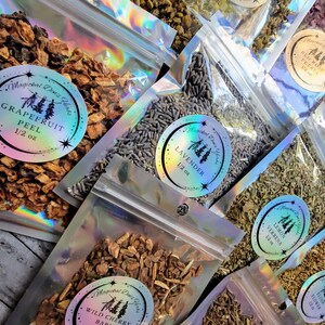 May include: A collection of small, clear plastic bags filled with dried herbs and flowers. Each bag has a label with the name of the herb and the weight. The herbs include grapefruit peel, lavender, lemon verbena, wild cherry bark, and a floral blend.