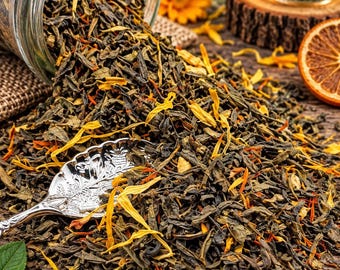 May include: A glass jar tipped over, spilling loose-leaf green tea mixed with dried yellow and orange flower petals. A silver-toned metal spoon with an ornate leaf-shaped bowl rests on the pile of tea leaves, accompanied by a cup of brewed tea.