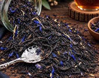 May include: A glass jar tipped over, spilling a blend of dark black tea leaves mixed with vibrant blue cornflower petals and small yellow flower pieces. A decorative silver spoon rests in the pile of loose tea, which is scattered across a dark wooden surface.