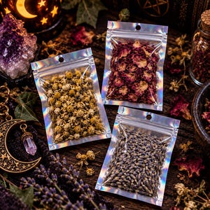 Op de afbeelding: Drie kleine, rechthoekige, holografische plastic zakjes gevuld met gedroogde planten. E&eacute;n zakje bevat gele kamillebloemen, &eacute;&eacute;n bevat donkerrode rozenblaadjes en &eacute;&eacute;n bevat kleine, gedroogde paarse lavendelknoppen.