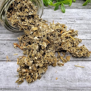 May include: Dried herbs spilling from a clear glass jar onto a gray wooden surface. The herbs are a mix of green and brown, with visible stems and leaves. Green foliage is in the background.