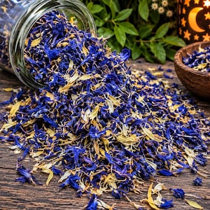 May include: A clear glass jar tipped on its side, spilling a mixture of dried blue cornflower petals and pale yellow botanical pieces onto a dark wooden surface. The dried floral blend is scattered across the wood, with a small wooden bowl nearby.