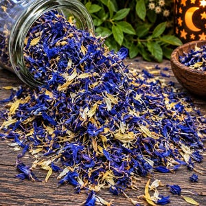 Op de afbeelding: Een glazen pot die op zijn kant ligt en een mengsel van gedroogde blauwe korenbloemblaadjes en lichtgele botanische stukjes morst op een donkerhouten oppervlak. De gedroogde bloemenmix ligt verspreid, met een kleine houten kom ernaast.