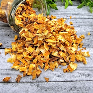 May include: A glass jar filled with dried orange peel pieces, spilling out onto a wooden surface. The orange peel pieces are a bright orange color and have a slightly rough texture.