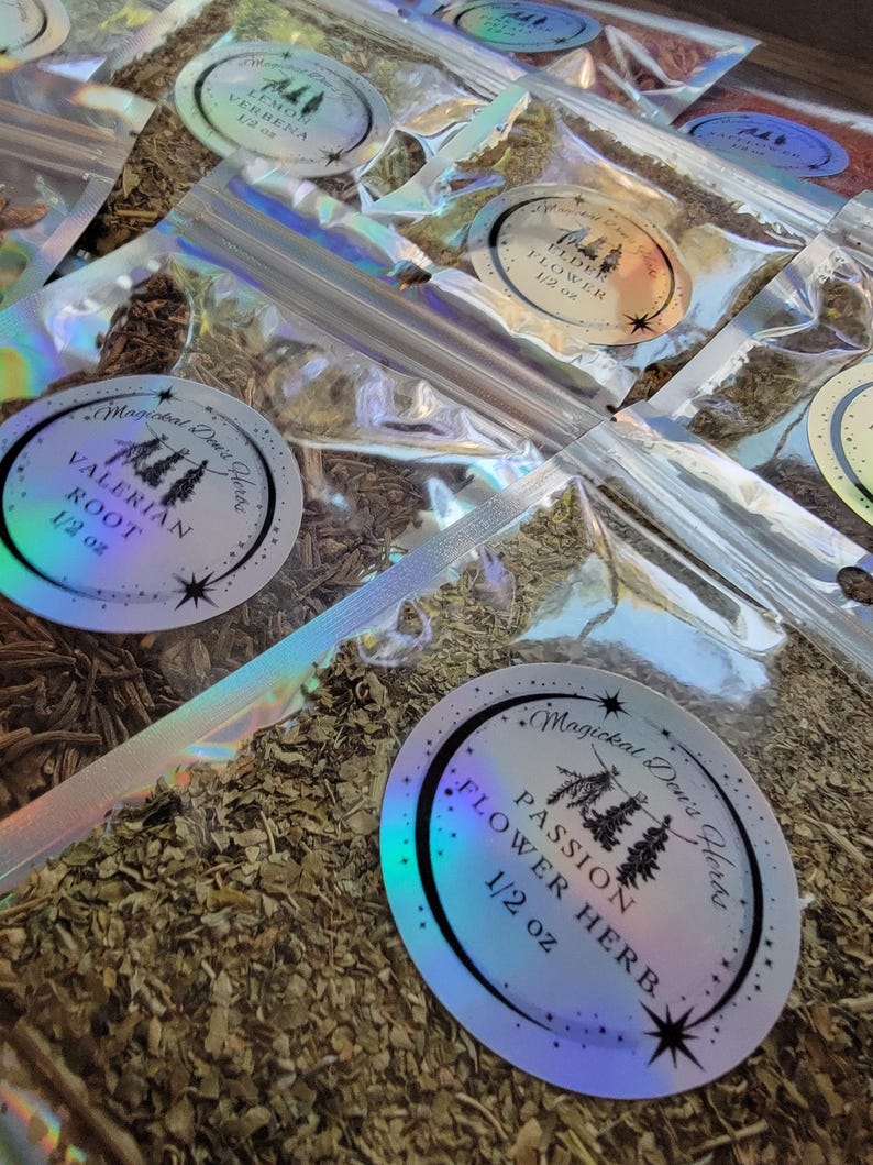 May include: A close-up of several small, clear plastic bags filled with dried herbs. Each bag has a round, iridescent sticker with a black and white illustration of a plant and the name of the herb, along with the text "Magickal Dew's Herbs" and the weight in ounces. The herbs include Lemon Verbena, Elder Flower, Valerian Root, and Passion Flower.