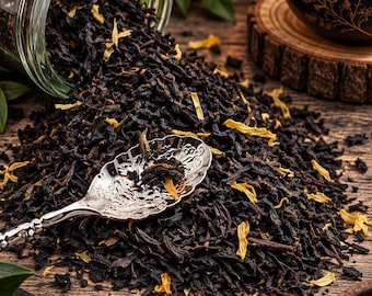 May include: A glass jar tipped over, spilling dark loose-leaf tea mixed with bright yellow flower petals onto a surface. A decorative silver spoon with an intricate embossed pattern rests in the center of the tea pile, surrounded by green leaves.