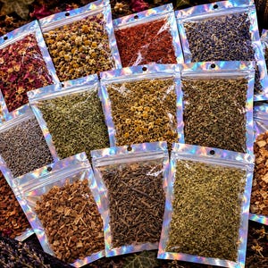 Op de afbeelding: Een verzameling kleine, doorzichtige holografische hersluitbare zakjes gevuld met diverse gedroogde kruiden, bloemen en botanische ingredi&euml;nten, waaronder rode rozenblaadjes, gele kamille, paarse lavendel, groene bladeren en bruine schors.