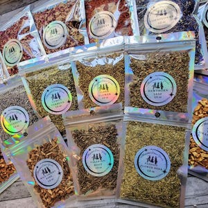 May include: Assortment of 12 small, clear, resealable bags filled with various dried herbs and flowers. Each bag has a label with the name of the herb or flower and the weight in ounces. The herbs and flowers are a variety of colors, including brown, green, white, and purple.