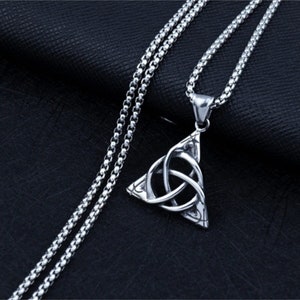 Könnte beinhalten: Eine silberfarbene Halskette mit einem dreieckigen Anhänger, der ein keltisches Knotenmuster zeigt. Der Anhänger hängt an einer feinen Kette. Die Halskette wird vor einem dunklen, strukturierten Hintergrund präsentiert.