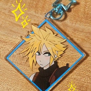 May include: A blue and white diamond-shaped acrylic keychain featuring a cartoon illustration of a blonde-haired character with a black turtleneck and a blue background. The keychain has a silver metal ring and a blue star charm.
