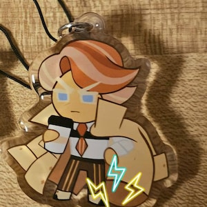 Op de afbeelding: Een transparante acryl sleutelhanger met een cartoonfiguur met oranje en roze haar, die een bruine jas, een wit shirt, een rode stropdas en een bruine broek draagt. Het personage houdt een witte beker vast en heeft blauwe ogen. De sleutelhanger heeft een zwart koord en een zilveren metalen ring. Het personage is omringd door gele bliksemschichten.
